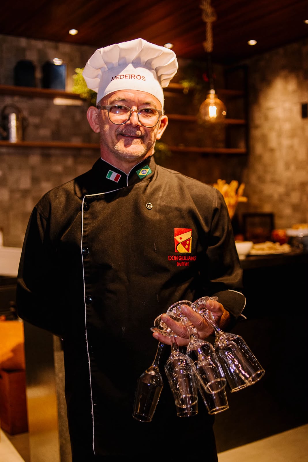 Chef de dólmã preto sorrindo, segurando taças