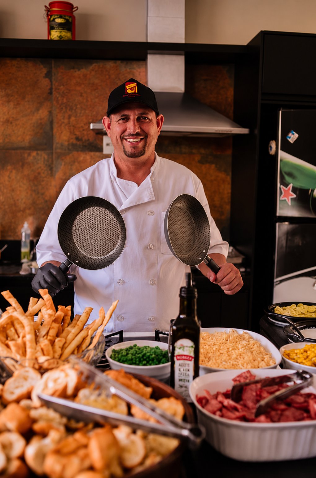 Chef sorrindo na estação de massas, segurando duas frigideiras, com ingredientes à frente